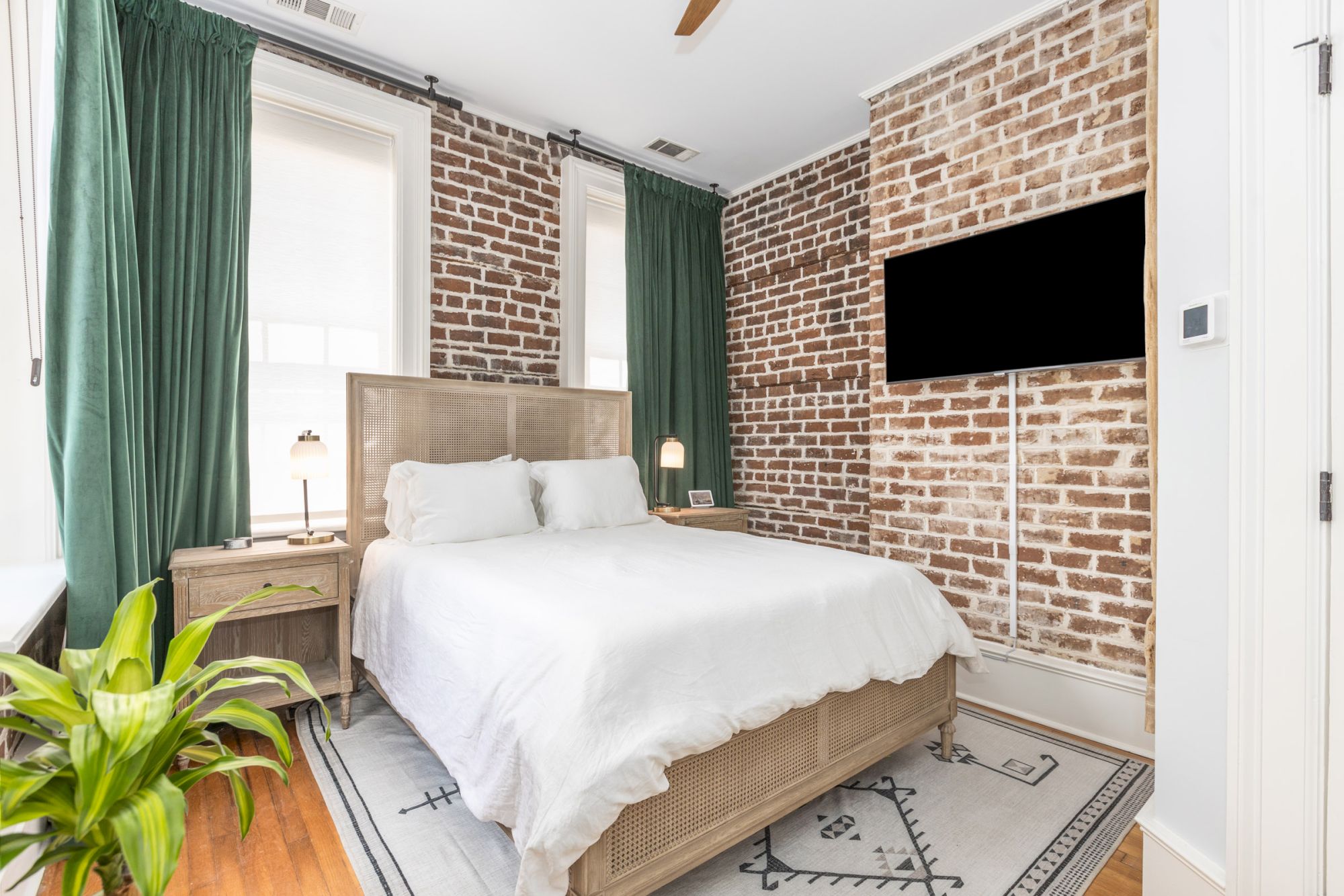 The image depicts a cozy bedroom featuring a bed with white linens, green curtains, a brick accent wall, a potted plant, and a wall-mounted TV.