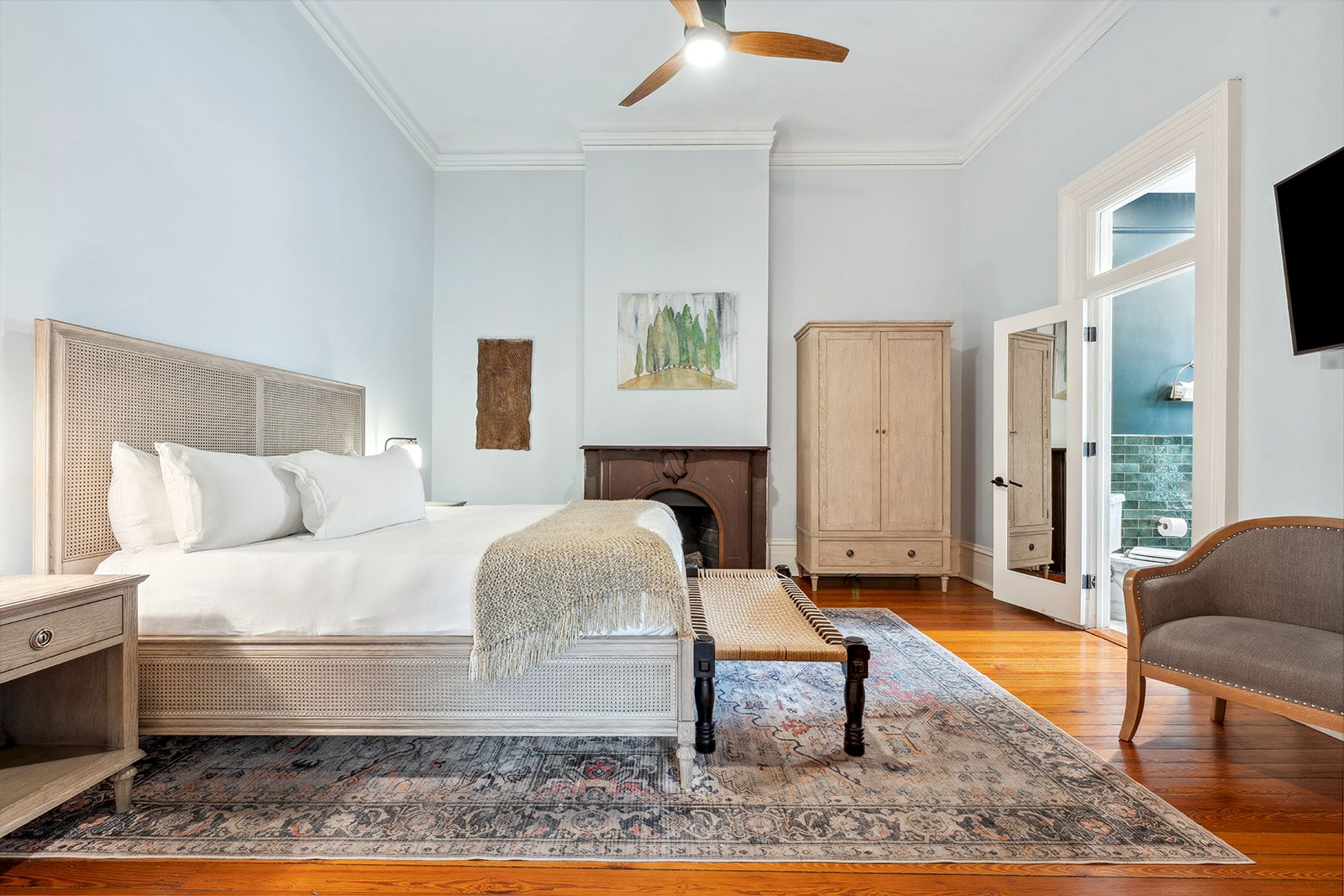 A bright, airy hotel-style bedroom with a king bed, ceiling fan, wooden floors, a rug, armoire, bench at foot, and a seating area. End of sentence.