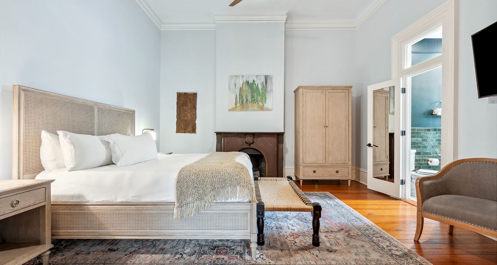 A bright, airy hotel-style bedroom with a king bed, ceiling fan, wooden floors, a rug, armoire, bench at foot, and a seating area. End of sentence.