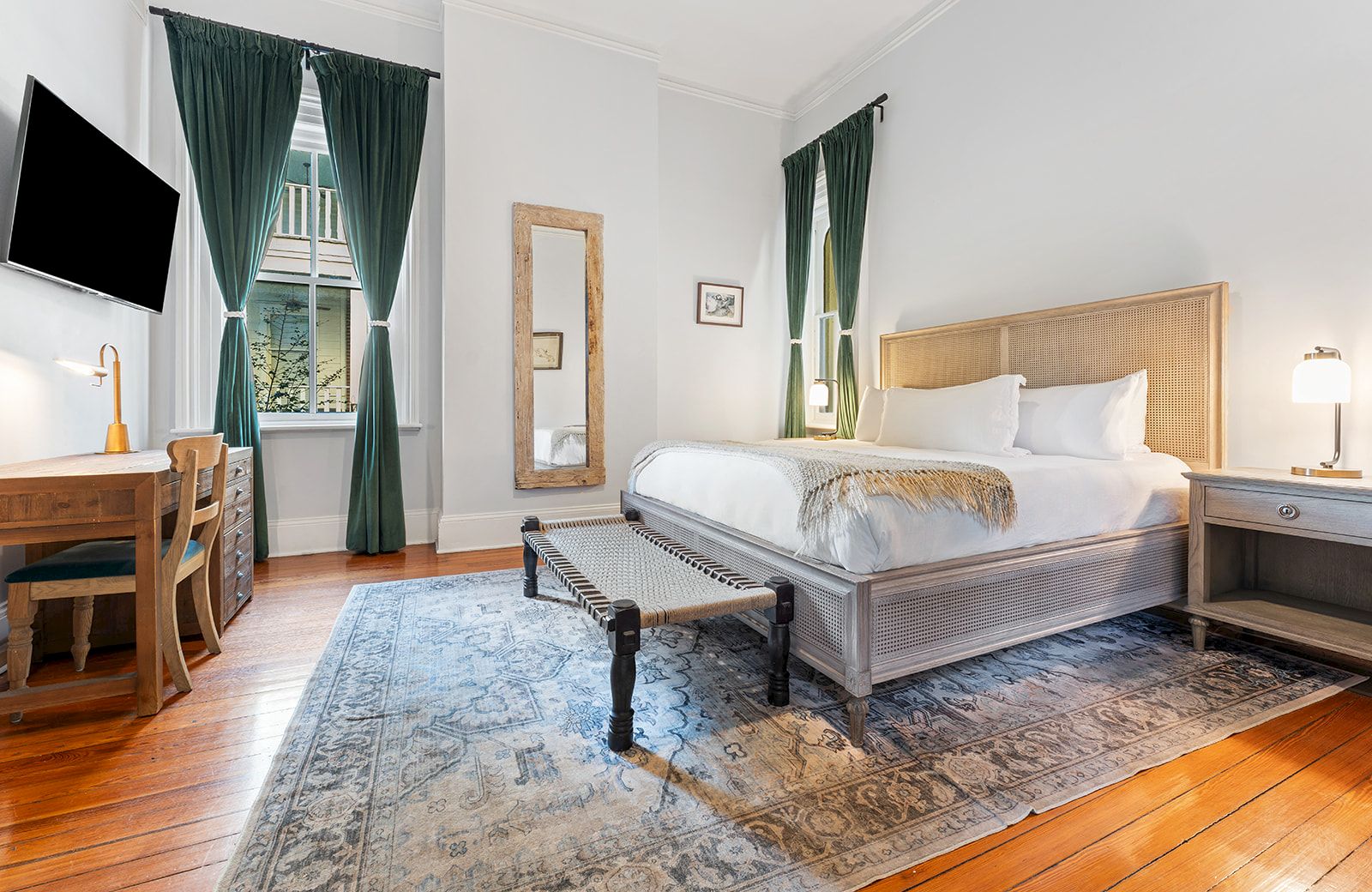 A bright hotel room with a large bed, side tables, a wooden floor, blue patterned rug, green curtains, a wall mirror, a desk with a chair, a TV, and soft bedside lighting.