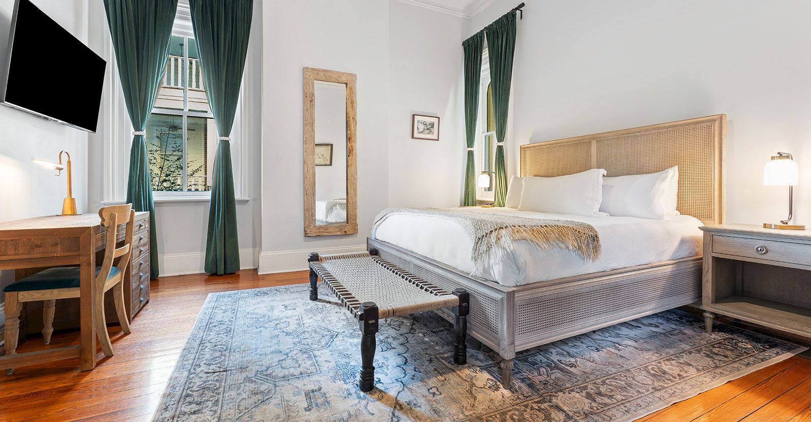 A bright hotel room with a large bed, side tables, a wooden floor, blue patterned rug, green curtains, a wall mirror, a desk with a chair, a TV, and soft bedside lighting.