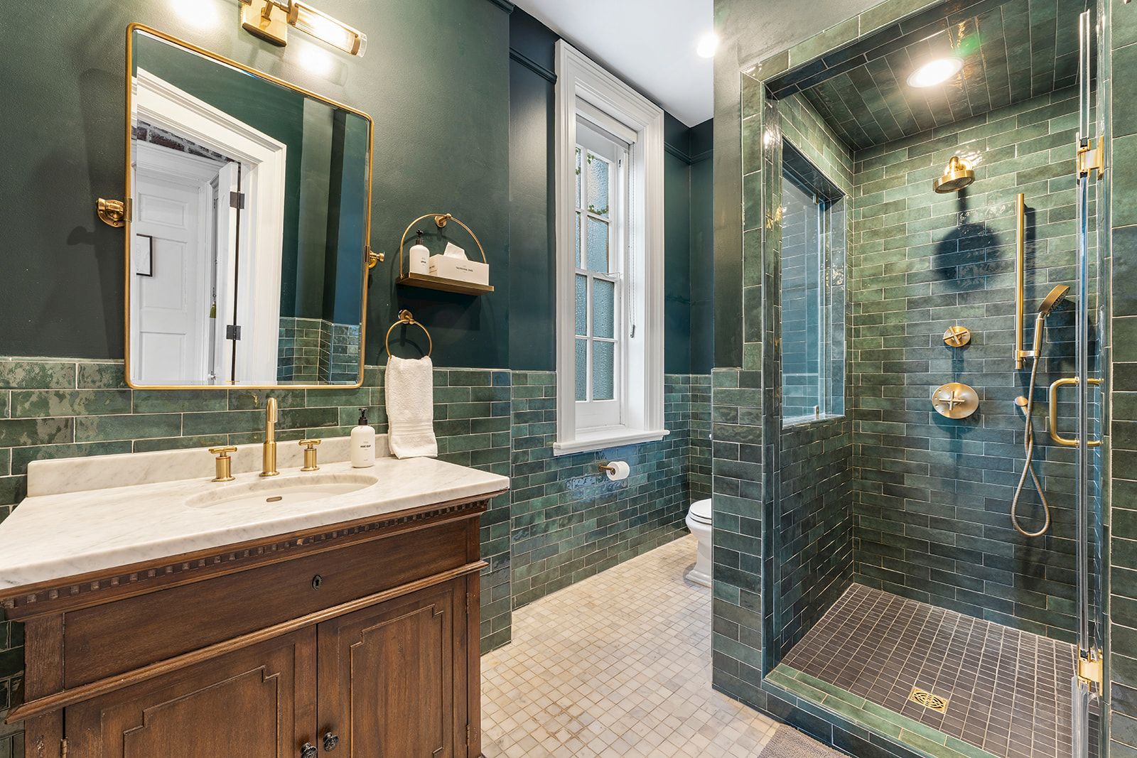 A green-tiled bathroom with a wooden vanity, gold fixtures, and a glass-enclosed shower. Top it at 140 characters, always ending the sentence.