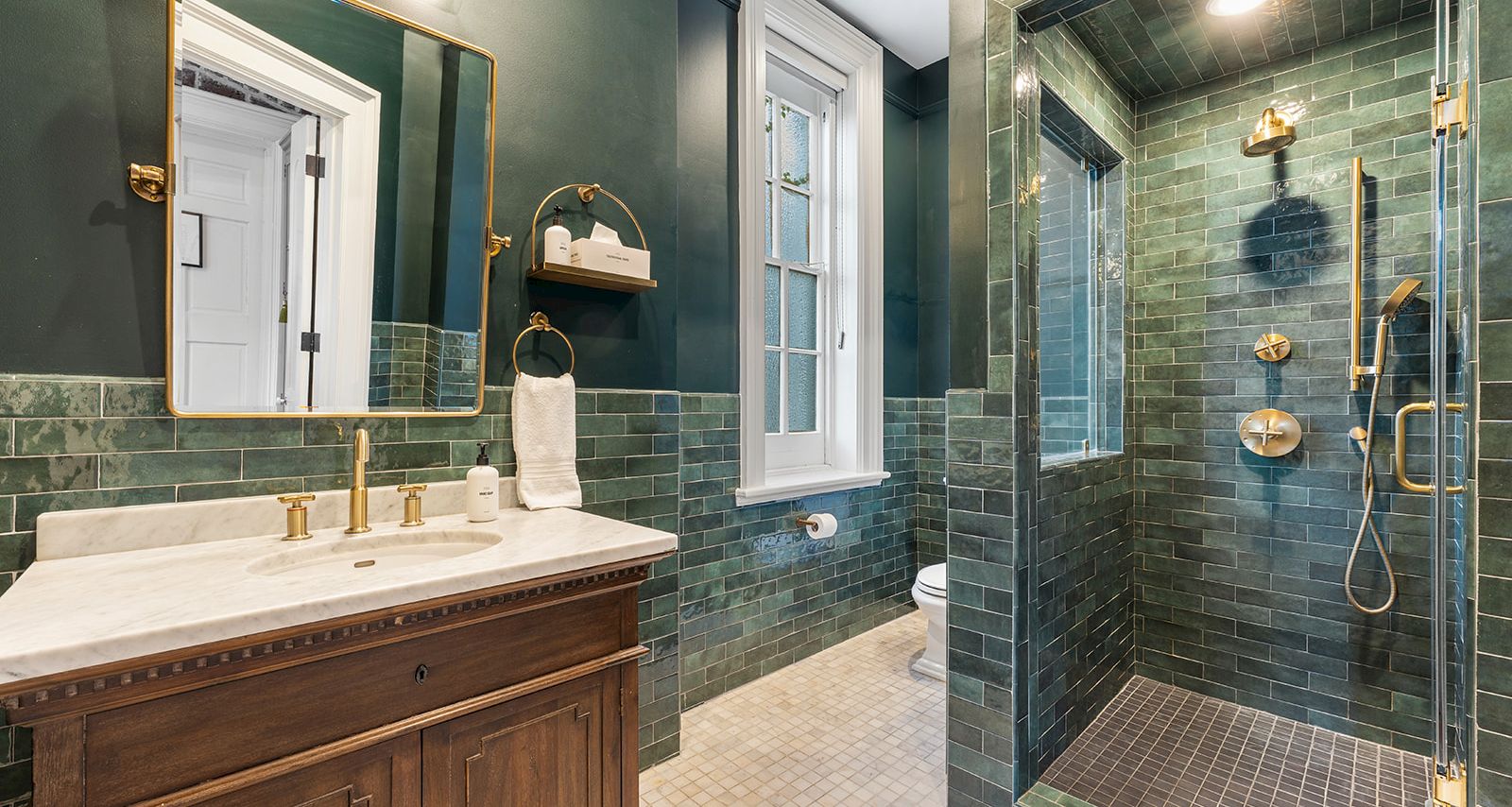 A green-tiled bathroom with a wooden vanity, gold fixtures, and a glass-enclosed shower. Top it at 140 characters, always ending the sentence.