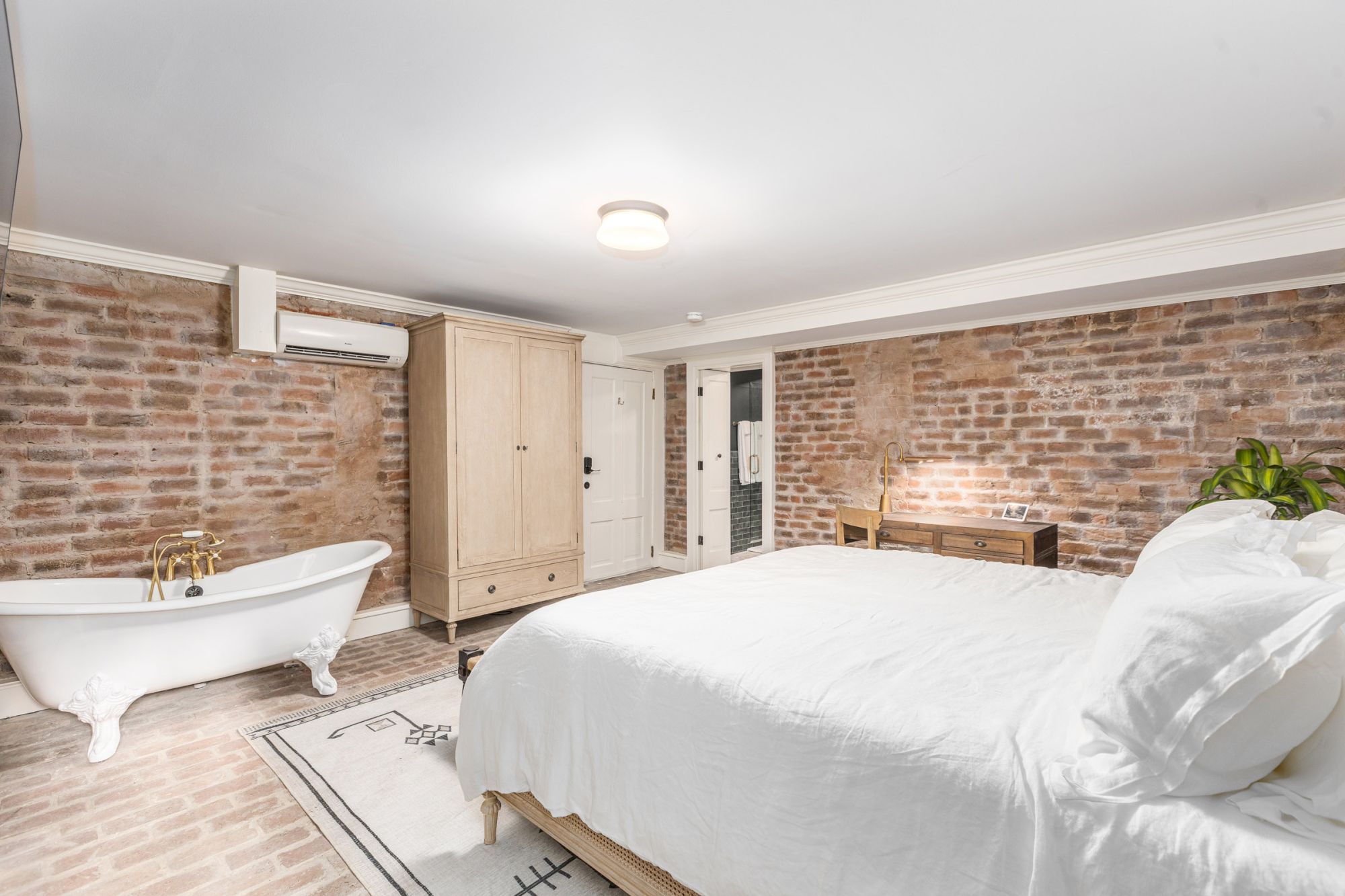 A cozy bedroom with exposed brick walls, featuring a bed, clawfoot bathtub, wooden wardrobe, small desk, and plants ending the sentence.