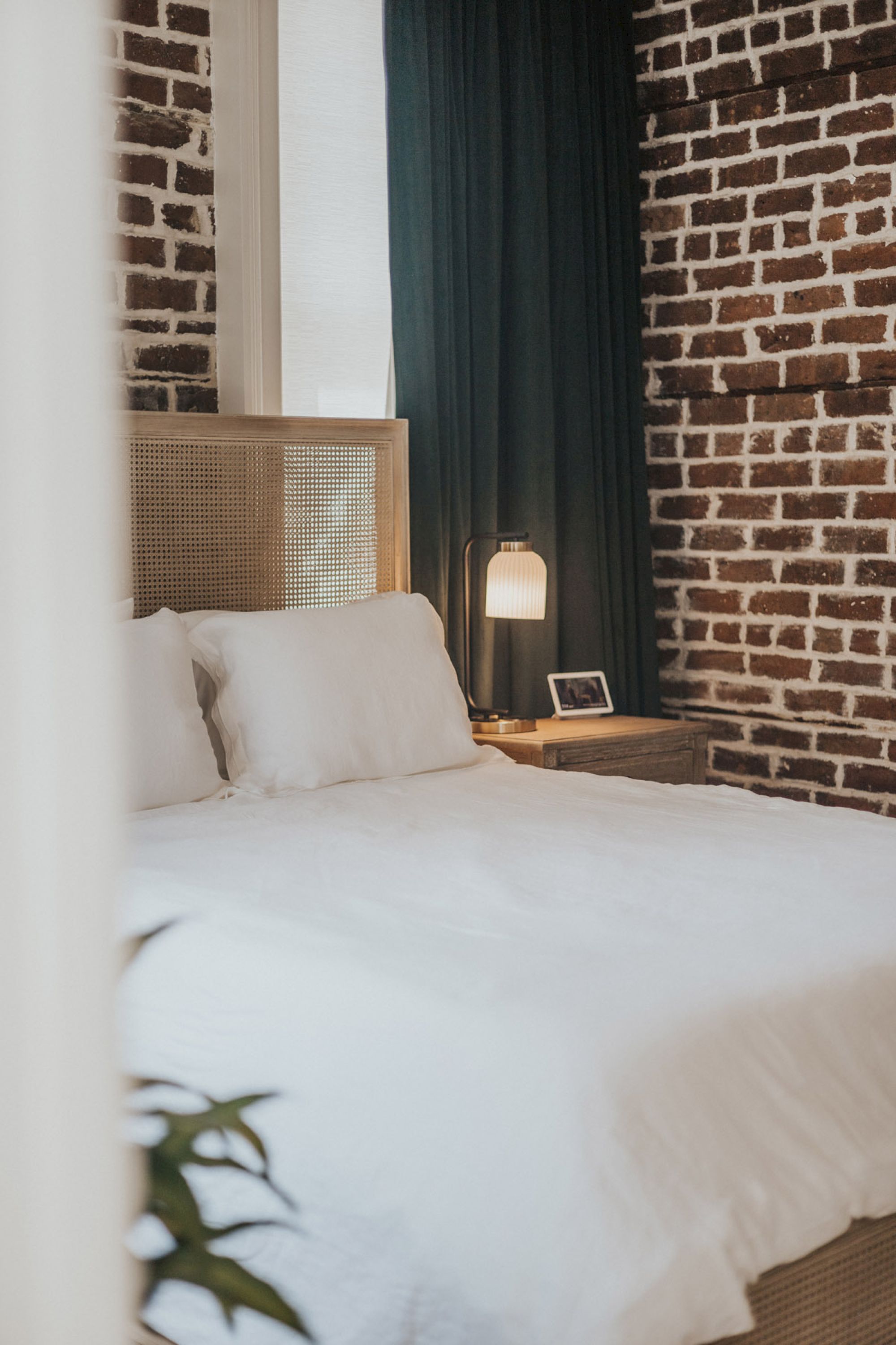 A cozy bedroom with a white duvet bed, a nightstand with a lamp, a small picture frame, and exposed brick walls.