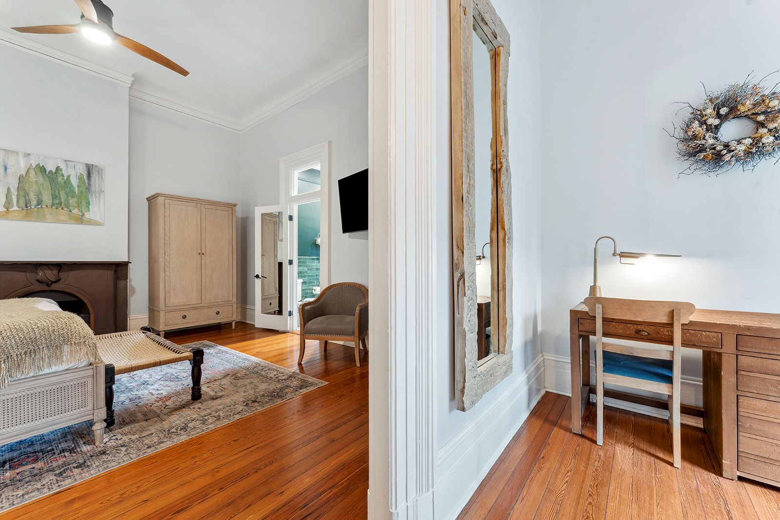 A bright hotel-style suite split by a doorway: left shows a wooden bed, wardrobe, rug, and seating in a pale blue-toned room; right features a wooden desk with a chair, lamp, and a decorative wreath on the wall.