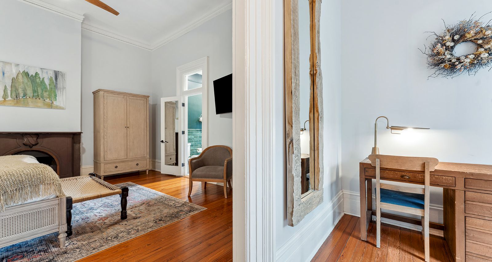 A bright hotel-style suite split by a doorway: left shows a wooden bed, wardrobe, rug, and seating in a pale blue-toned room; right features a wooden desk with a chair, lamp, and a decorative wreath on the wall.