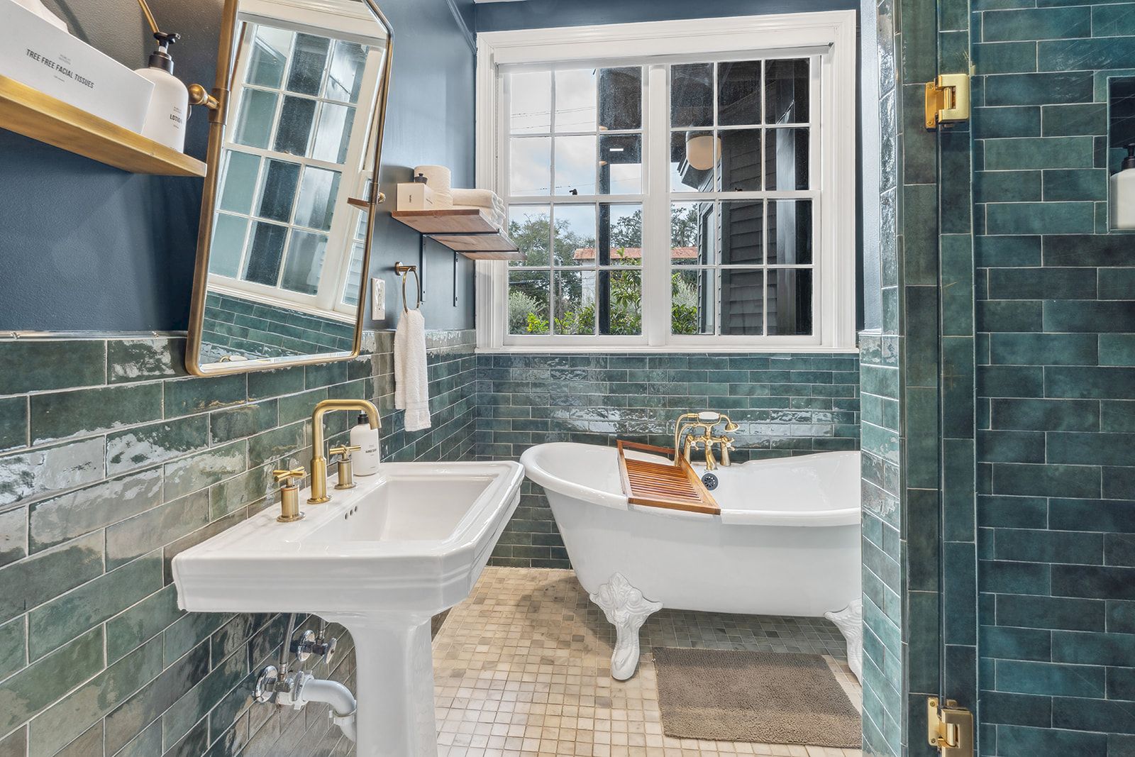 A vintage-style bathroom with a freestanding clawfoot tub, pedestal sink, green tile walls, window, and brass fixtures, cozy and classic.
