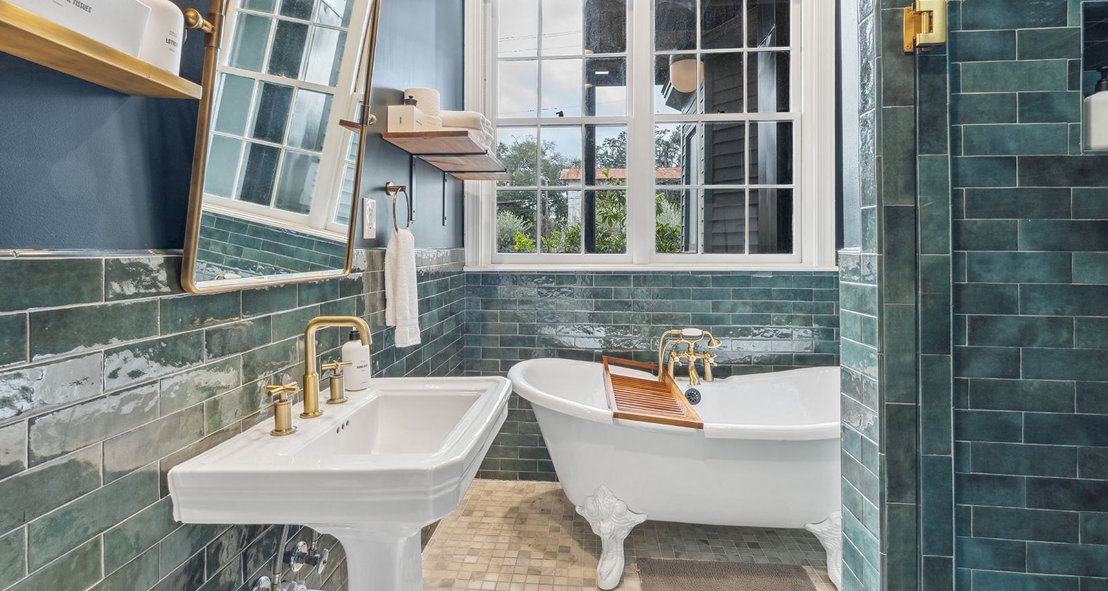A vintage-style bathroom with a freestanding clawfoot tub, pedestal sink, green tile walls, window, and brass fixtures, cozy and classic.