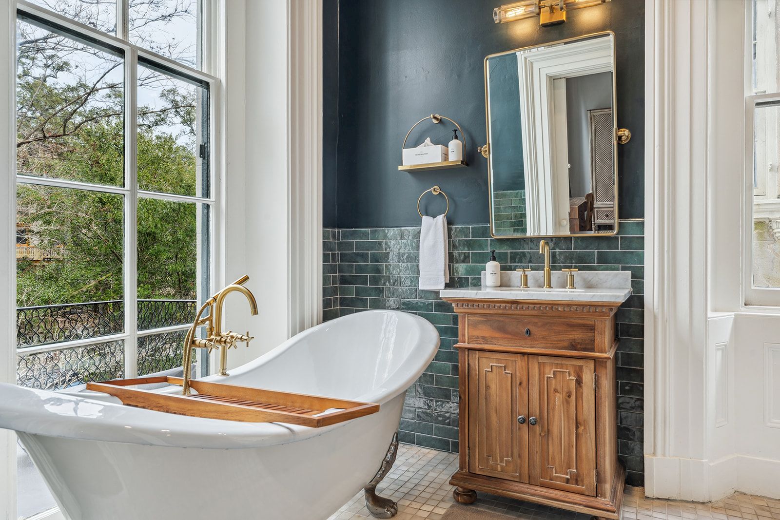 A freestanding clawfoot tub by a large window, wooden bath caddy, brass faucet, navy tiled wall, small wooden vanity, and a mirror with soft lighting.