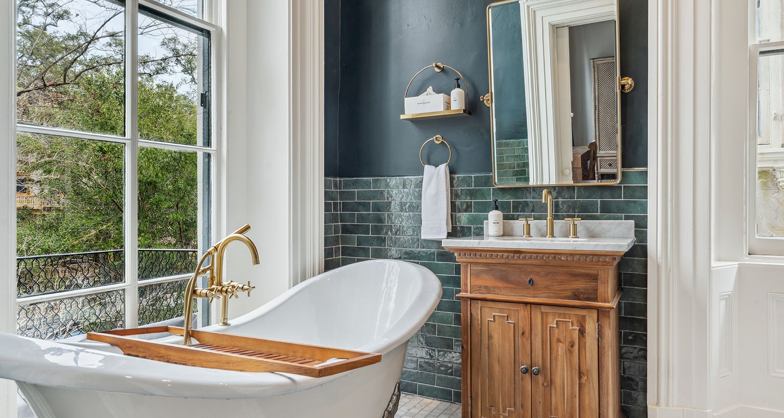 A freestanding clawfoot tub by a large window, wooden bath caddy, brass faucet, navy tiled wall, small wooden vanity, and a mirror with soft lighting.