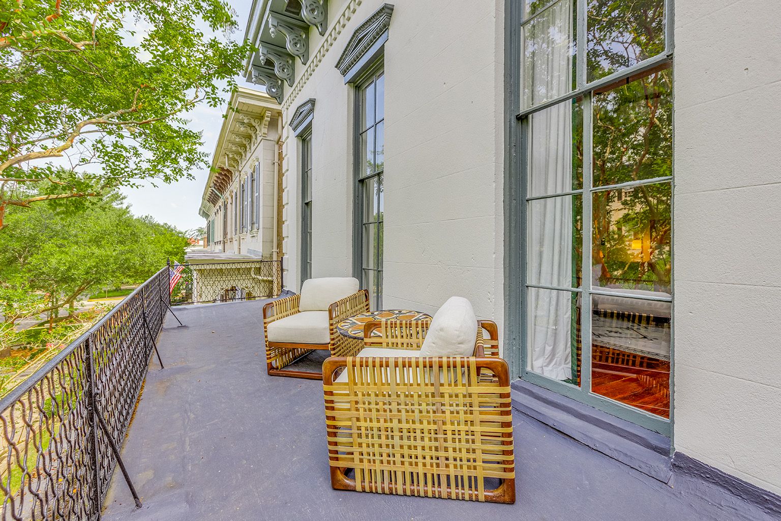 A spacious apartment balcony with wicker chairs, patterned cushions, tall windows, and leafy trees beside an elegant historic building.