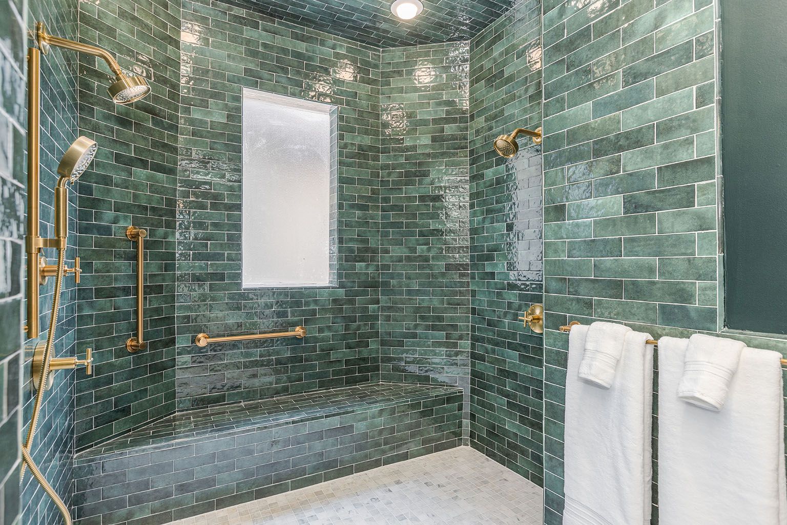 A modern tiled shower with green wall tiles, gold showerheads, a bench, and folded white towels along the right wall.