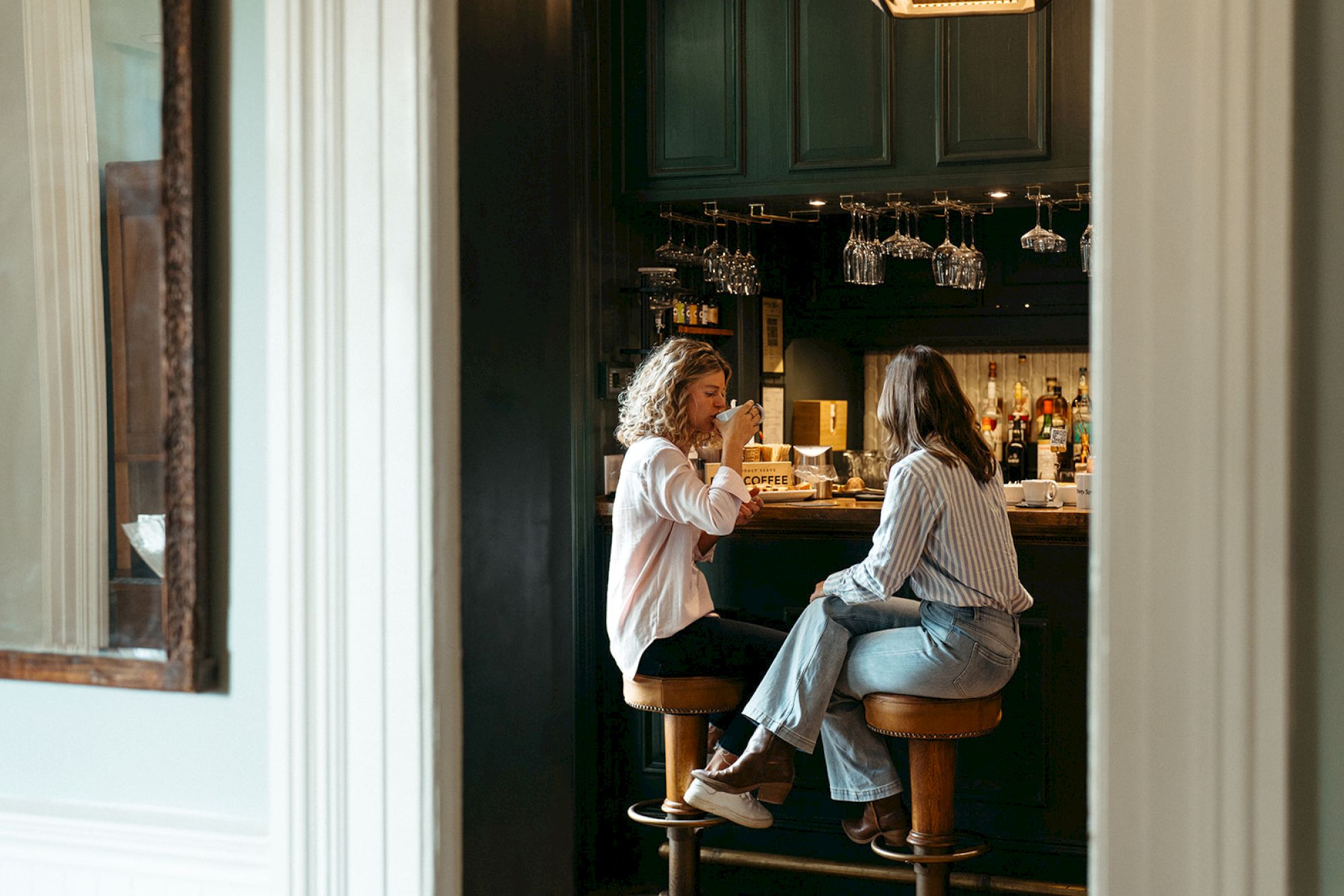 Two people are seated at a bar, engaged in conversation, with shelves of bottles and glasses in the background.