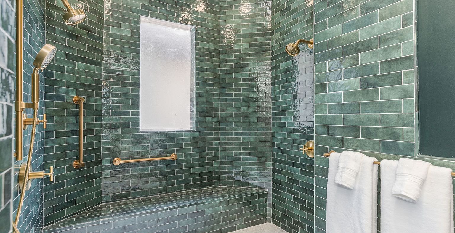 A green-tiled accessible shower area with multiple showerheads, a built-in bench, grab bars, a window, and two white towels hanging nearby.