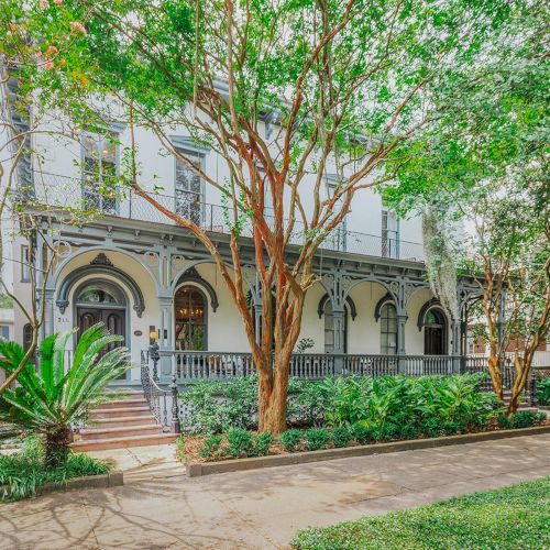 The image shows a charming house with large windows, surrounded by lush trees and plants, and a stone pathway leading to the entrance.