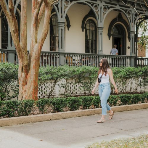 A woman wearing sunglasses and casual clothes walks on a sidewalk, in front of a historic-looking building with elaborate architecture and lush greenery.