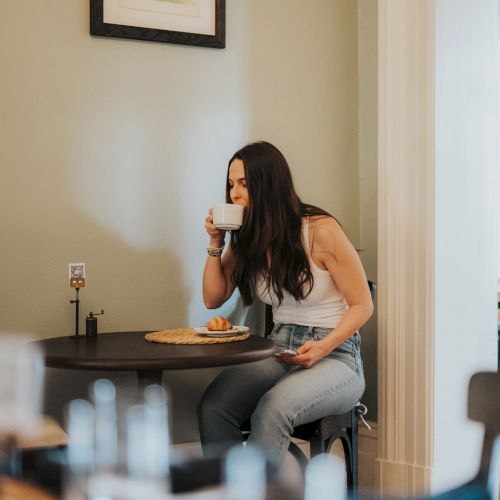 A woman is sitting at a table in a cozy setting, drinking from a cup with a plate of food in front of her and holding a phone.