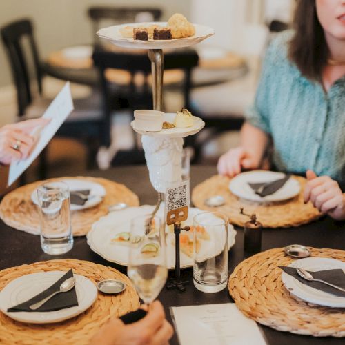 Three people sit around a table with plates, glasses, and a tiered stand of food. One person holds a menu, and another a glass of water.