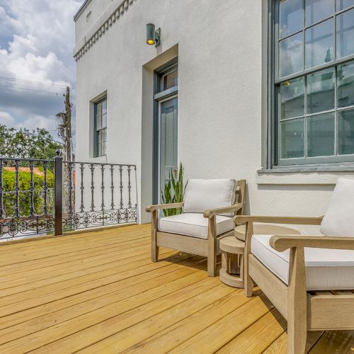 A sunlit balcony with two cushioned wooden chairs, a potted plant, and an ornate iron railing against a bright sky.