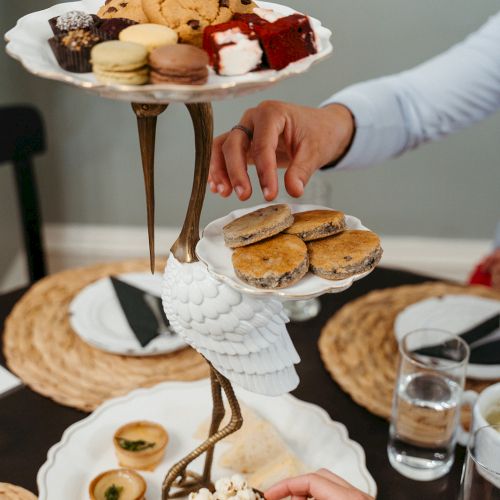 A multi-tier dessert tray with pastries and cookies, topped with a hand reaching to grab a treat, against a pale background.