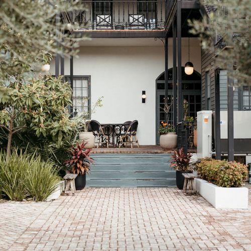 A narrow alley with stone walls, potted plants, and a wooden gate leading to a doorway, flanked by ivy-covered arches and a cozy courtyard.