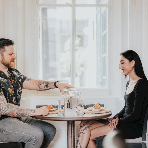 Two women sit at a table in a bright kitchen, chatting and sharing a coffee or tea.