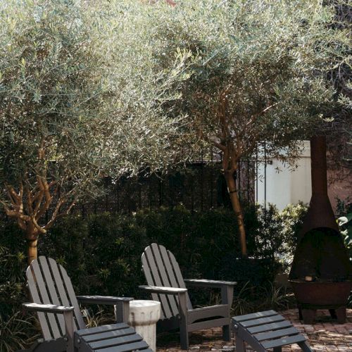 A sunny courtyard patio with two wooden rocking chairs, an outdoor fireplace, and olive trees and shrubs.