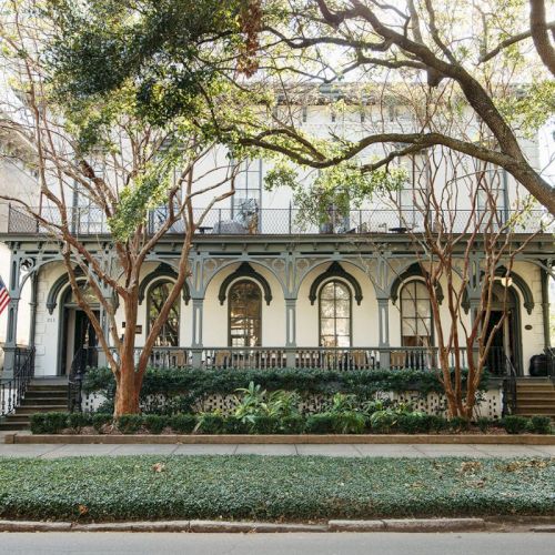 A sunny courtyard with leafy trees and shrubs in front of a white building featuring arched windows and a central staircase.