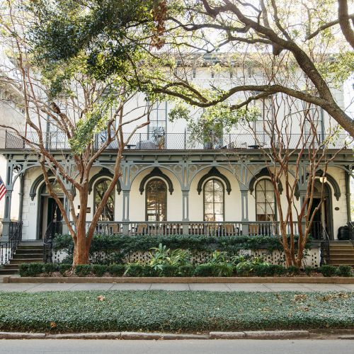 A stately colonial-style building with a long colonnade and arched windows, set among green trees and a manicured lawn.