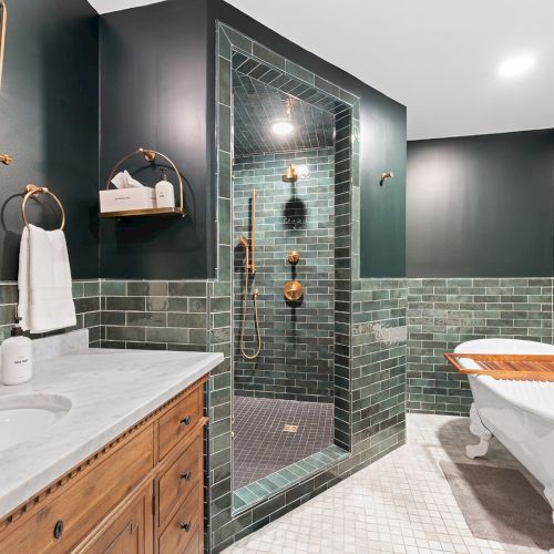 A modern bathroom with a glass-enclosed shower, teal-green tiled walls, a wooden vanity, a freestanding tub, and brass fixtures, stylish lighting.