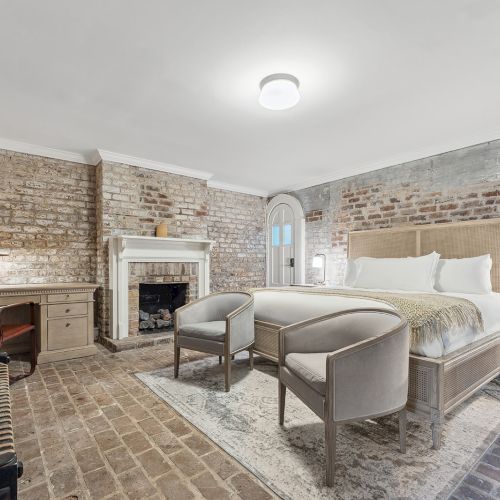 A stylish hotel-like bedroom with an exposed brick wall, a plush king bed, two chairs at the foot, a cozy fireplace, and soft modern lighting.