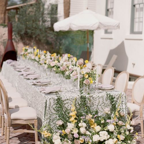 A wedding cake table setup with tiered cake, floral arrangements, and chairs outdoors.