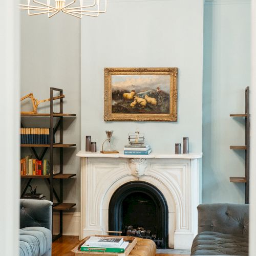 A cozy living room with a pale blue wall, white mantel and fireplace, a brown leather sofa, a framed painting above the mantel, and a bookshelf beside the fireplace.