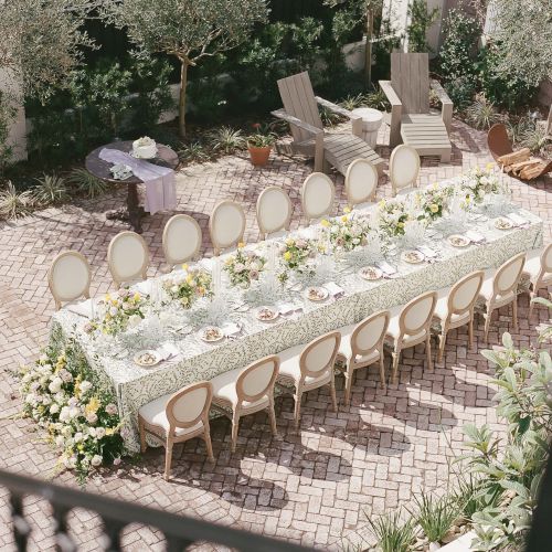 A long outdoor table set with chairs, white tablecloth, and place settings in a garden or courtyard.