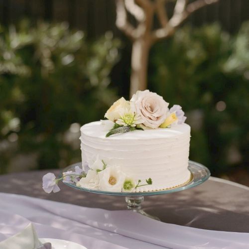 A white wedding cake topped with pale flowers sits on a glass stand outdoors in a garden setting.