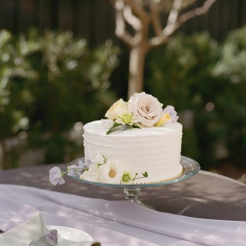 A small white frosted cake with pink and yellow flower decorations sits on a wooden surface, placed on a light gray cake stand.
