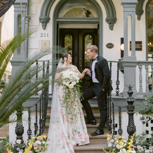 A bride in a lace gown and veil stands with a groom or officiant inside a decorated doorway, surrounded by floral arrangements and tropical plants, indoors.