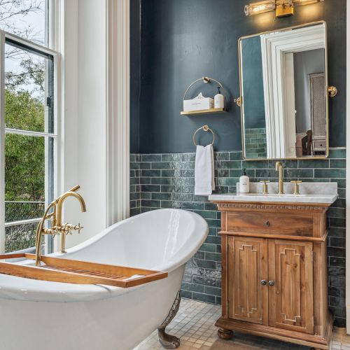A luxurious bathroom with a freestanding tub by a large window, a wooden vanity, blue tile walls, and brass fixtures, bathed in natural light.