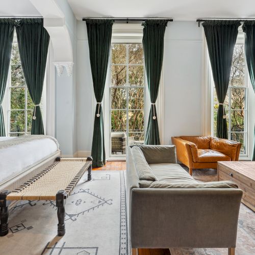A bright hotel-style room with a large bed, dark draped windows, a brown sofa and gray chairs, wooden coffee table, and a light rug.