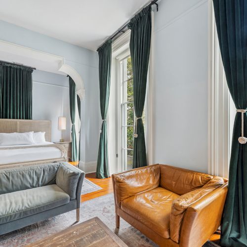 A stylish hotel room with a large bed, a gray sofa, a tan leather armchair, dark green drapes, and floor-to-ceiling windows letting in natural light.