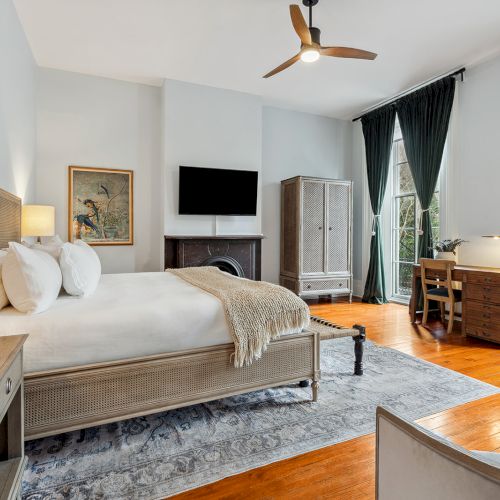 A luxurious hotel room with a king bed, fireplace, flat-screen TV, wooden furniture, large windows with dark green curtains, and a workspace.