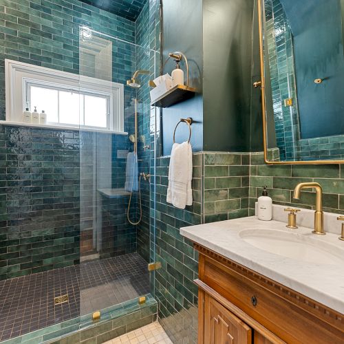 A modern green-tiled bathroom with a glass shower, gold fixtures, and a wooden vanity with a marble countertop, all in a compact space.