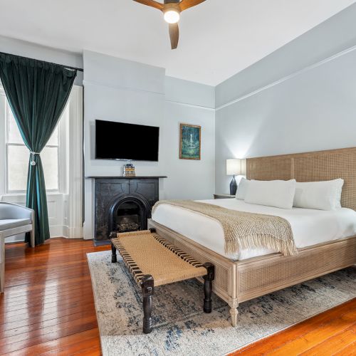 A bright hotel bedroom with a wooden floor, king bed, fireplace, a wall-mounted TV, green curtains, and a seating area by the window.