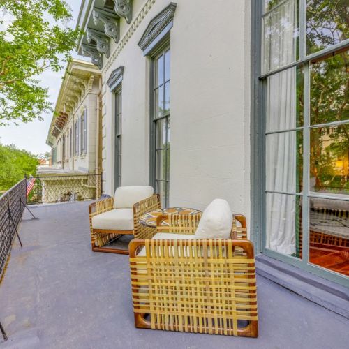 A spacious apartment porch with wicker chairs, an American flag, and tall windows beside lush green trees.