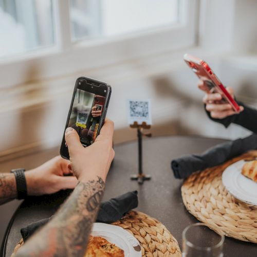A person is enjoying a meal with a phone displaying a colorful photo, while a latte, pastry, and small plate sit on a wooden table.