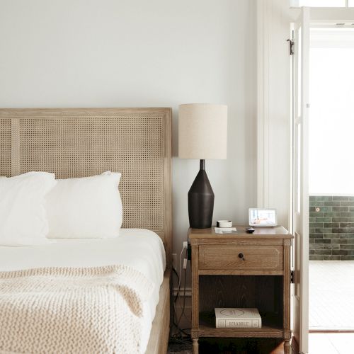 A cozy bedroom with a light wood headboard, white bedding, a bedside table, lamp, and a bottle with a glass on a polished dark floor.