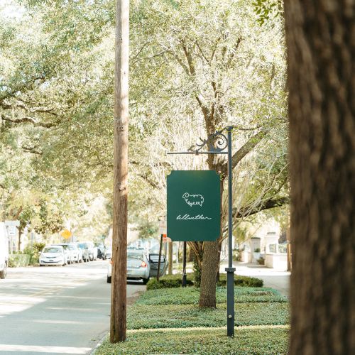 A tree-lined campus path with a green sign on a post reading &ldquo;Campus Green&rdquo; near a bench and walkway, sunny day.