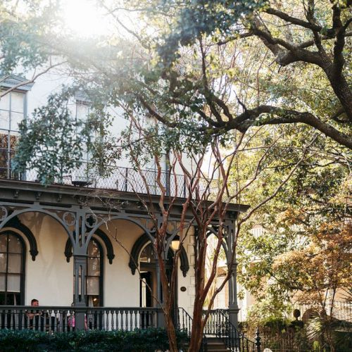 Sunlit courtyard with a white building&rsquo;s arched porch, leafy trees, and a walkway bordered by shrubs and railings.