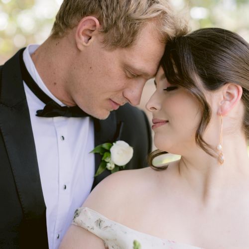 A wedding couple in formal attire, a man in a tuxedo and a woman in a wedding dress sharing a tender moment outdoors.